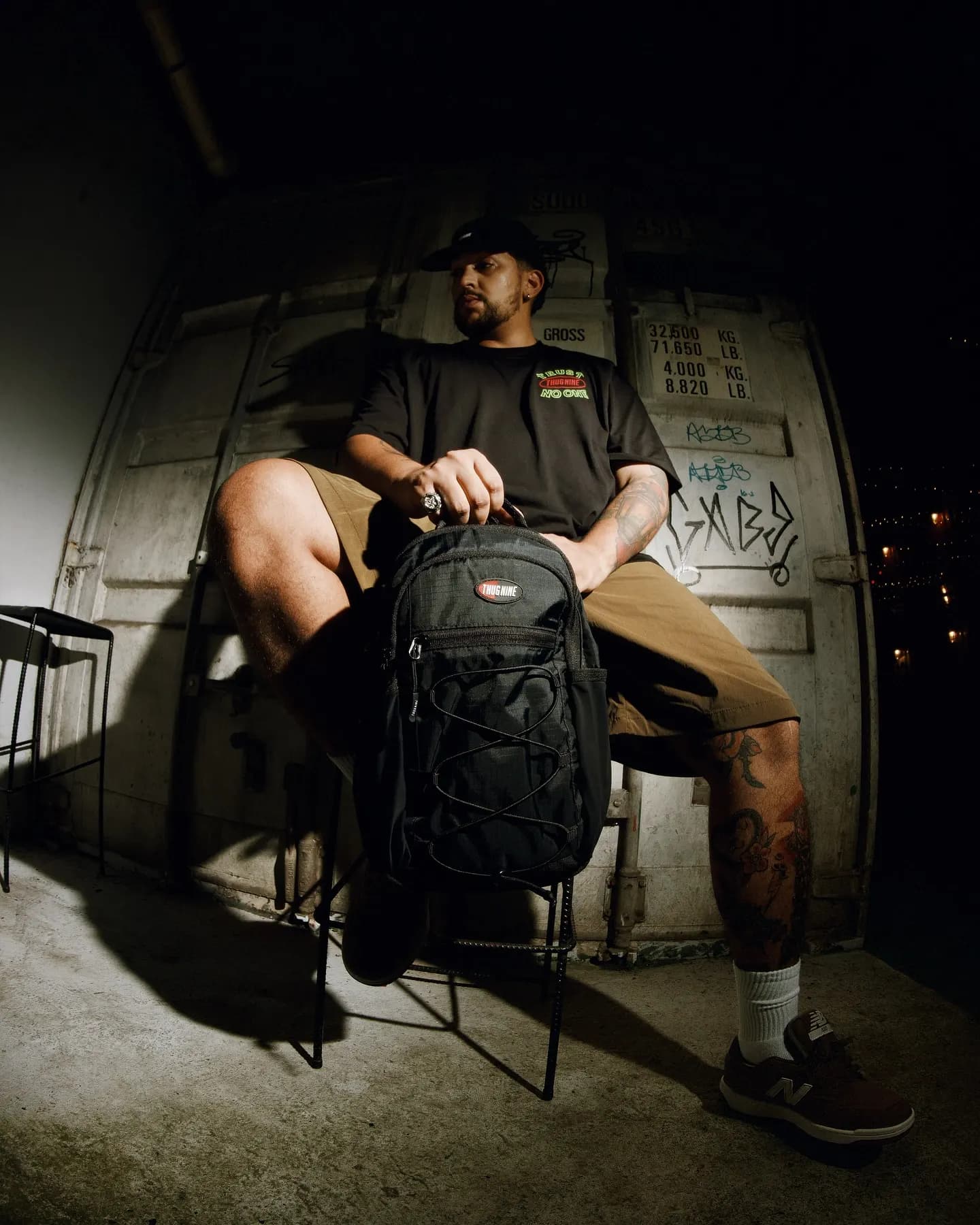 Rapaz sentado em frente à porta do container, com a mochila preta da Thug Nine no colo